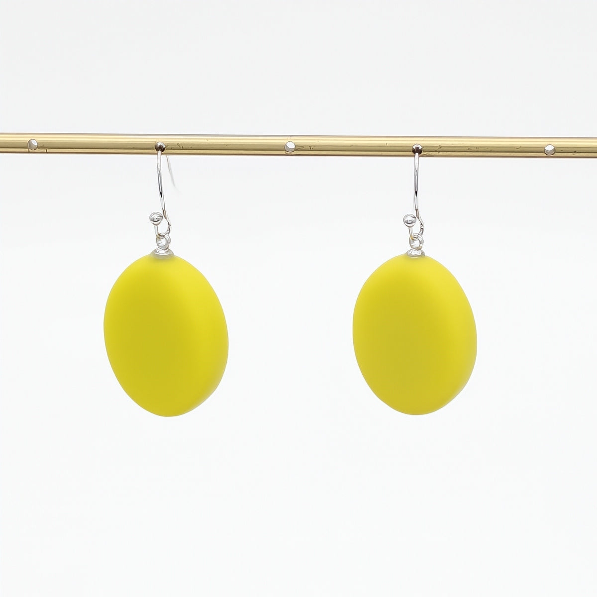 A pair of bright yellow oval drop earrings with silver fishhook wires, hanging from a gold-colored horizontal display bar against a plain white background.