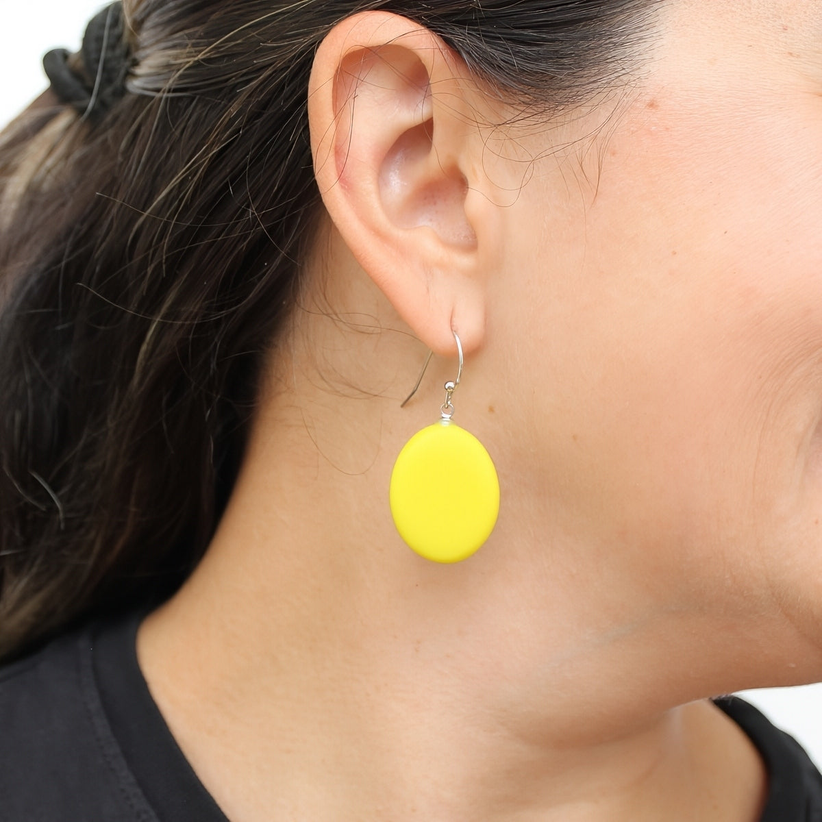 A close-up of a woman's ear wearing a bright yellow oval drop earring with a silver fishhook wire.