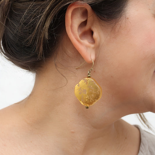 A close-up of a woman's ear and neck wearing a single irregular gold-yellow dangle earring with a gold-tone hook.