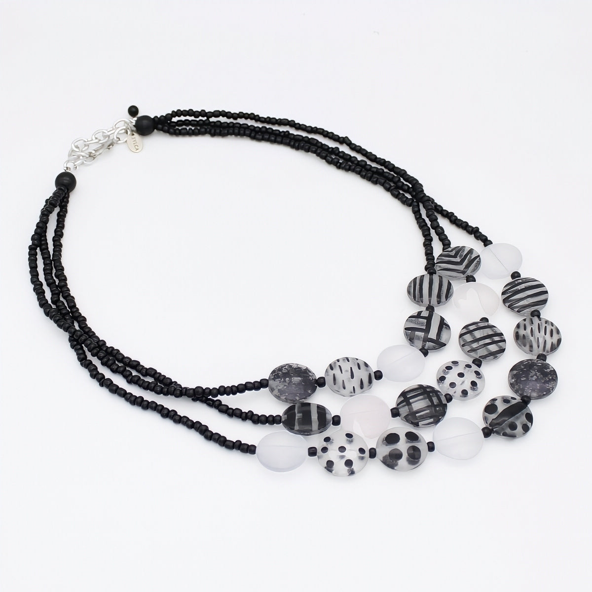 A multi-strand statement necklace featuring black beaded strands, translucent white beads, and patterned black-and-white disc beads with stripes, dots, and marbled textures, finished with a silver-tone clasp.