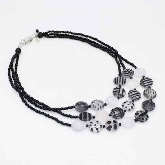 A multi-strand statement necklace featuring black beaded strands, translucent white beads, and patterned black-and-white disc beads with stripes, dots, and marbled textures, finished with a silver-tone clasp.