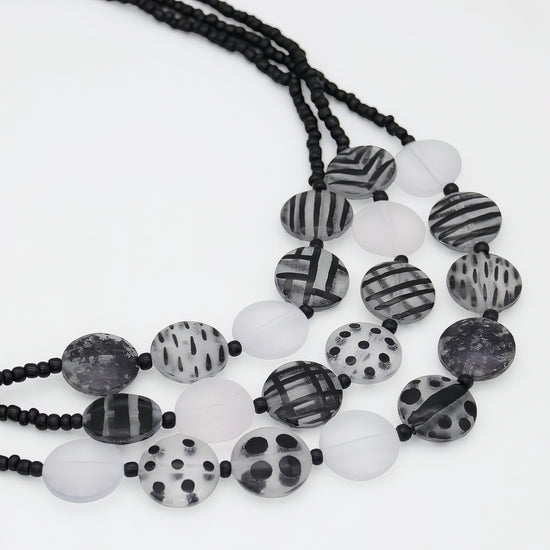 A multi-strand beaded necklace featuring black and white patterned round beads, translucent white beads, and black cord, displayed on a clean white background.