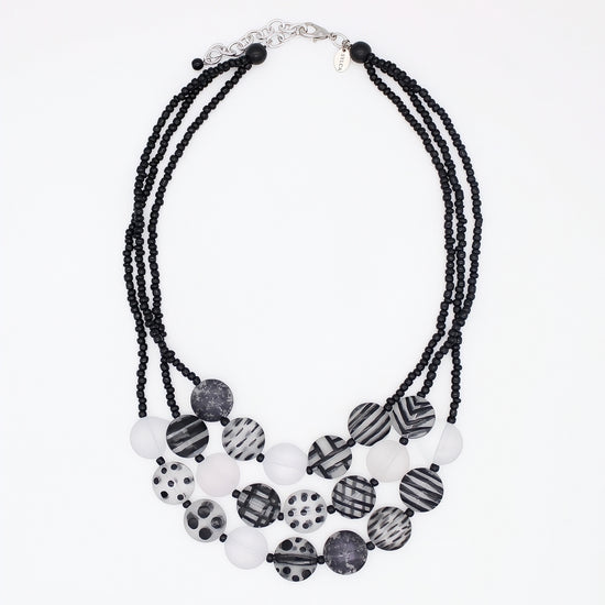 A multi-strand statement necklace featuring black beaded cords and a tiered arrangement of round beads in black, white, and gray patterns, including stripes, polka dots, and marbled textures, on a plain white background.