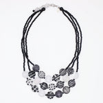A multi-strand statement necklace featuring black beaded cords and a tiered arrangement of round beads in black, white, and gray patterns, including stripes, polka dots, and marbled textures, on a plain white background.