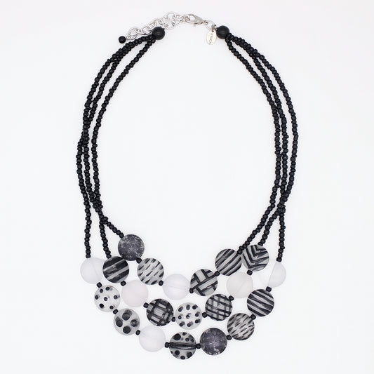A multi-strand statement necklace featuring black beaded cords and a tiered arrangement of round beads in black, white, and gray patterns, including stripes, polka dots, and marbled textures, on a plain white background.