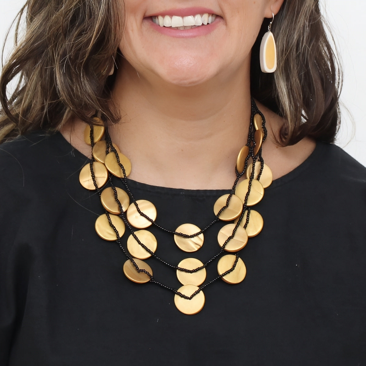 A close-up of a woman wearing a multi-strand statement necklace with flat gold-tone disc charms on black cords, paired with a black top and a matching gold drop earring.