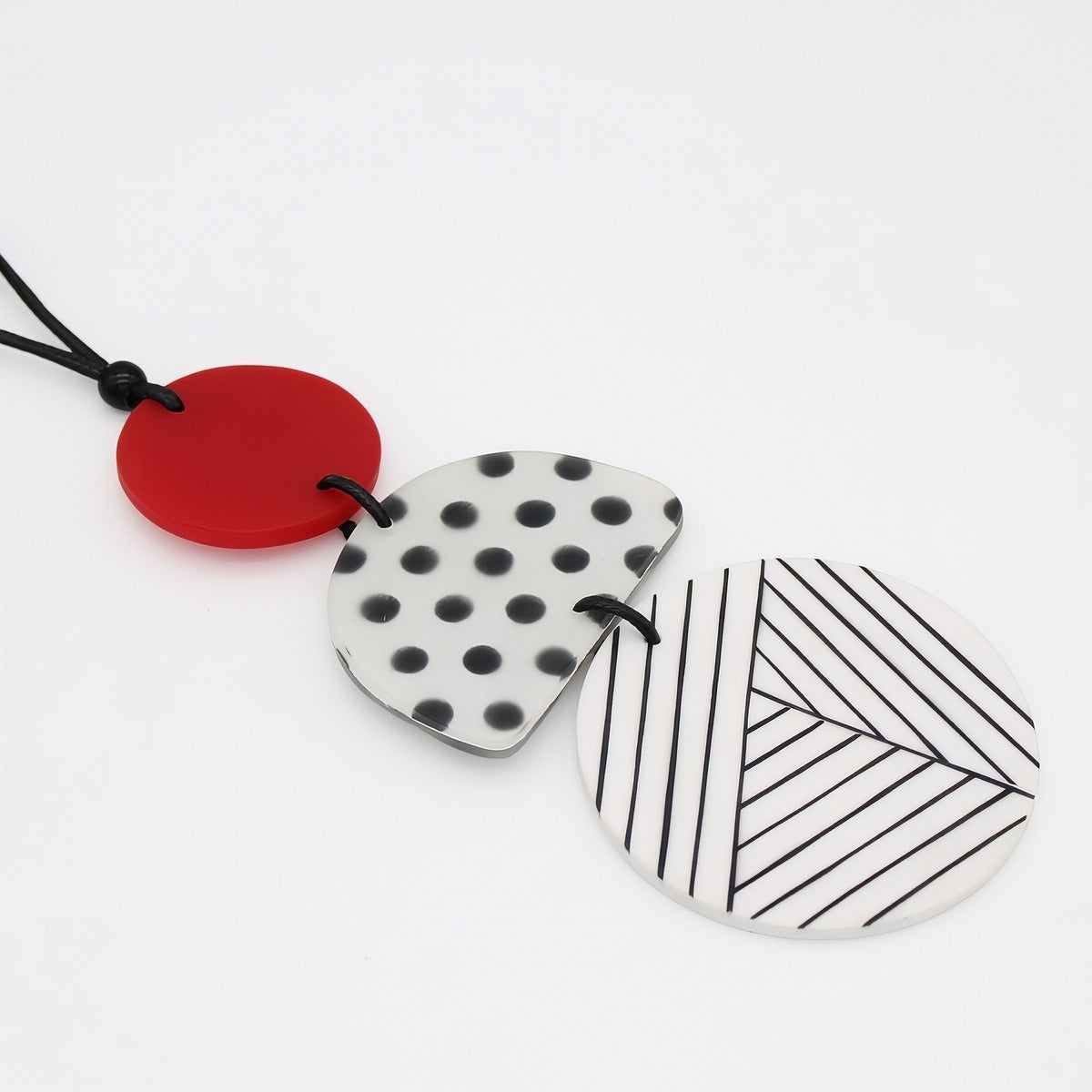 Three circular pendant necklaces with black cords, featuring a solid red pendant, a white pendant with black polka dots, and a white pendant with black diagonal stripes, arranged on a clean white background.