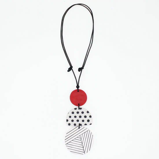 A modern multi-piece pendant necklace featuring a black cord, a solid red disc, a white disc with black polka dots, and a white disc with black diagonal lines, shown on a plain white background.