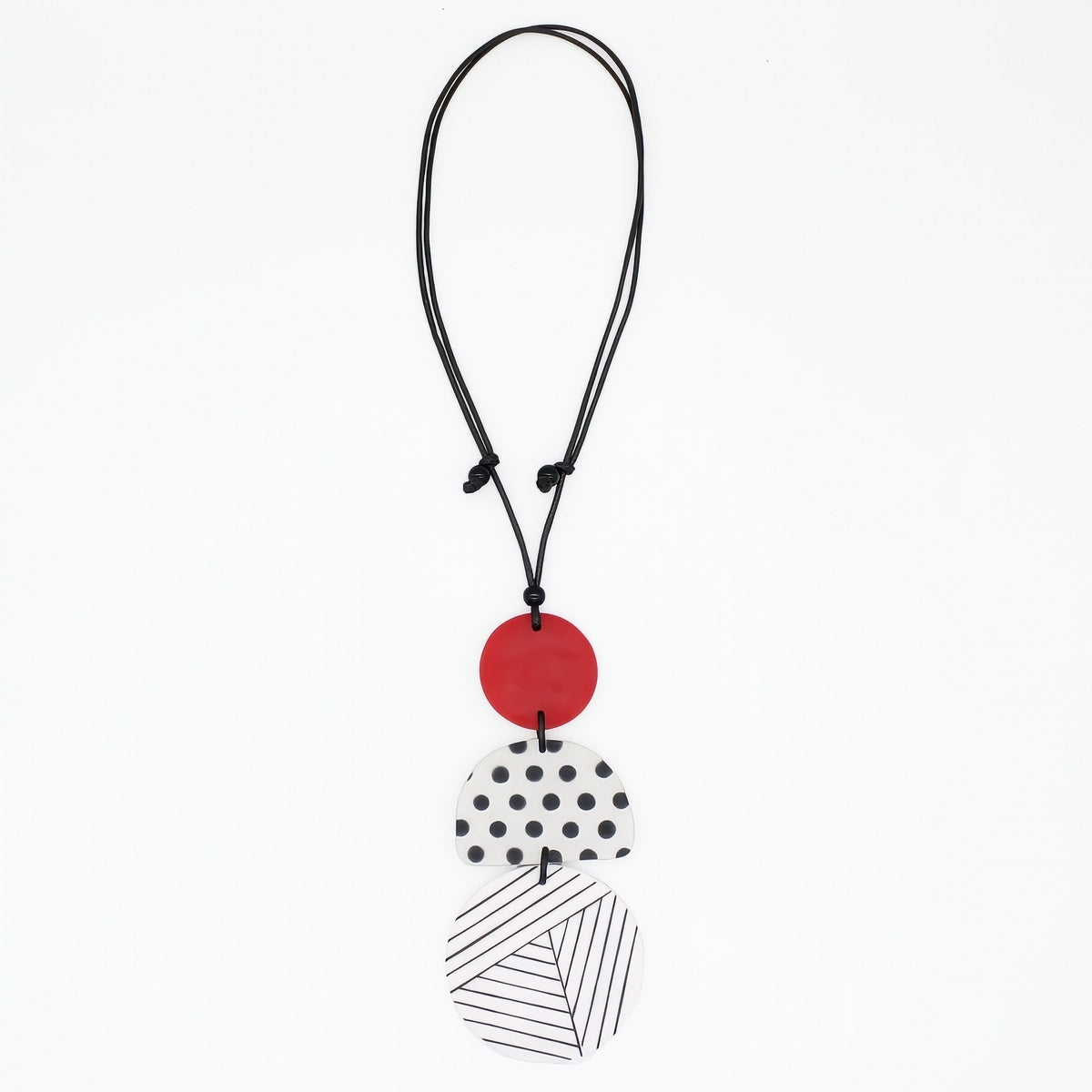 A modern multi-piece pendant necklace featuring a black cord, a solid red disc, a white disc with black polka dots, and a white disc with black diagonal lines, shown on a plain white background.