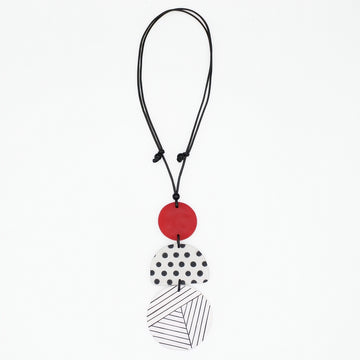 A modern multi-piece pendant necklace featuring a black cord, a solid red disc, a white disc with black polka dots, and a white disc with black diagonal lines, shown on a plain white background.