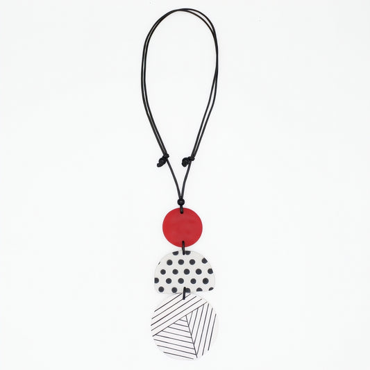 A modern multi-piece pendant necklace featuring a black cord, a solid red disc, a white disc with black polka dots, and a white disc with black diagonal lines, shown on a plain white background.