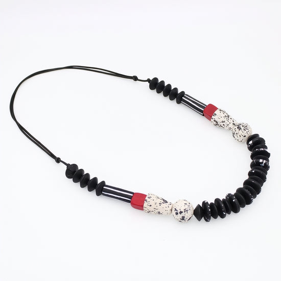 A beaded necklace featuring black disc beads, striped black and white beads, and two red accent beads with white speckled center beads, displayed on a clean white background.