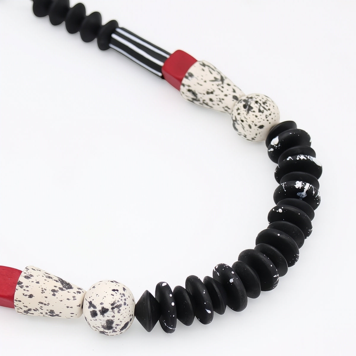 A close-up of a beaded necklace featuring matte black disc beads, speckled cream and white beads, and red accent bands on a plain white background.