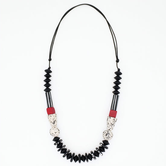 A beaded necklace featuring black faceted beads, black and white striped sections, textured silver-toned details, and small red accent beads, suspended on a black cord against a plain white background.