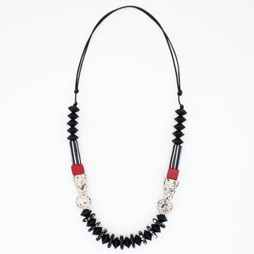 A beaded necklace featuring black faceted beads, black and white striped sections, textured silver-toned details, and small red accent beads, suspended on a black cord against a plain white background.