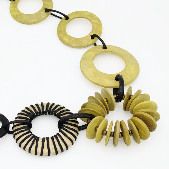 A close-up of a chunky beaded necklace featuring olive green stone disc links, a striped black and beige cord ring, and a cluster of small olive green disc beads, all threaded on black cords against a white background.