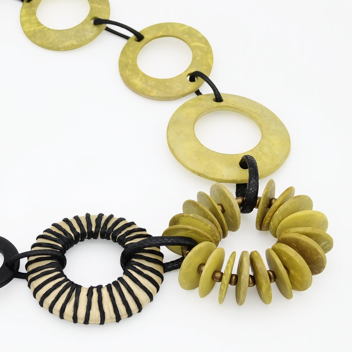 A close-up of a chunky beaded necklace featuring olive green stone disc links, a striped black and beige cord ring, and a cluster of small olive green disc beads, all threaded on black cords against a white background.