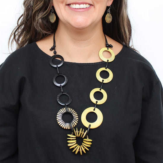 A close-up of a woman wearing a statement necklace with alternating black and gold circular and sunburst links, paired with matching gold earrings and a black top.