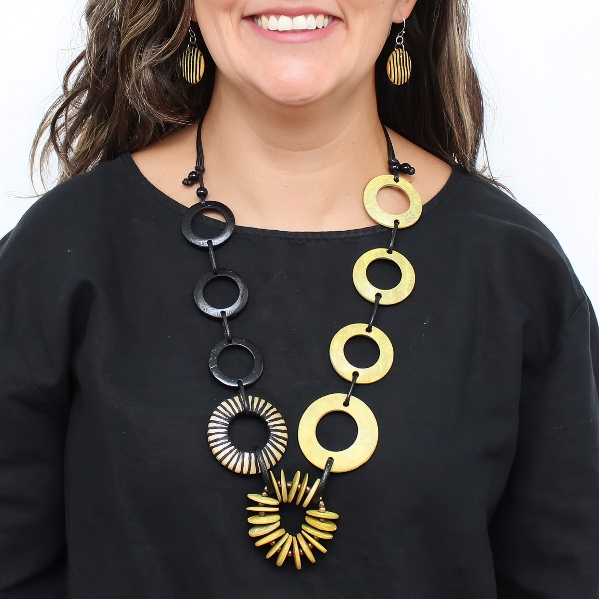 A close-up of a woman wearing a statement necklace with alternating black and gold circular and sunburst links, paired with matching gold earrings and a black top.