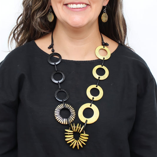 A close-up of a woman wearing a statement necklace with alternating black and gold circular and sunburst links, paired with matching gold earrings and a black top.