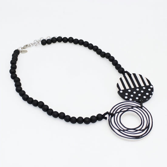 A black beaded necklace with two unique geometric charms: one heart-shaped charm featuring black and white stripes and white polka dots, and another circular ring charm wrapped in black and white striped cord, displayed on a clean white background.