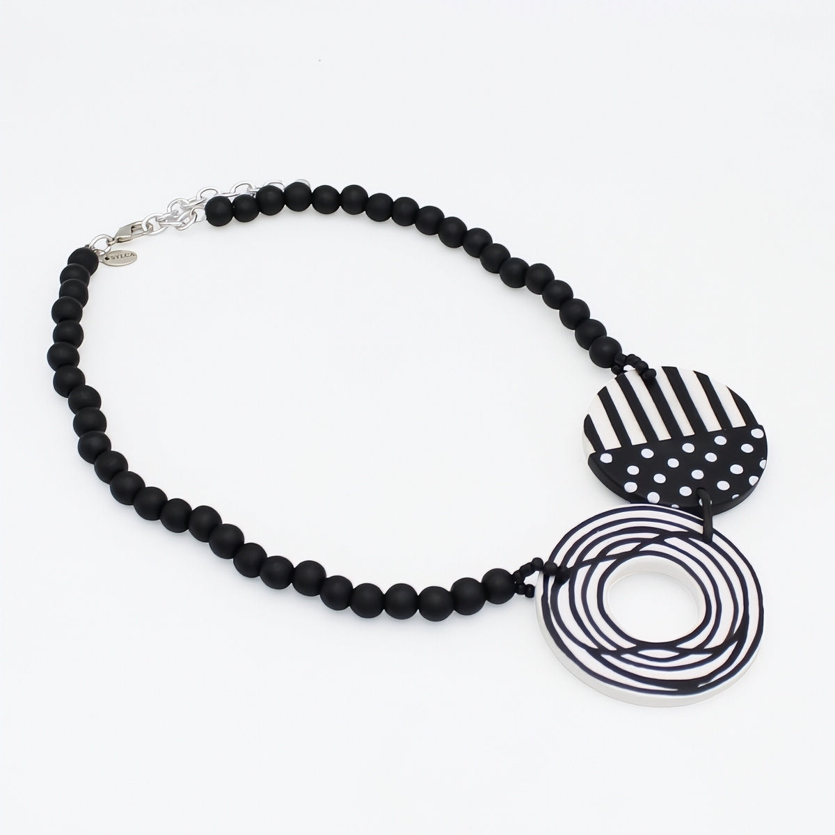 A black beaded necklace with two unique geometric charms: one heart-shaped charm featuring black and white stripes and white polka dots, and another circular ring charm wrapped in black and white striped cord, displayed on a clean white background.