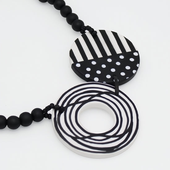 Black and white geometric pendant necklace featuring a striped and polka-dot circular pendant and a coiled ring pendant, suspended over a black beaded chain on a white background.