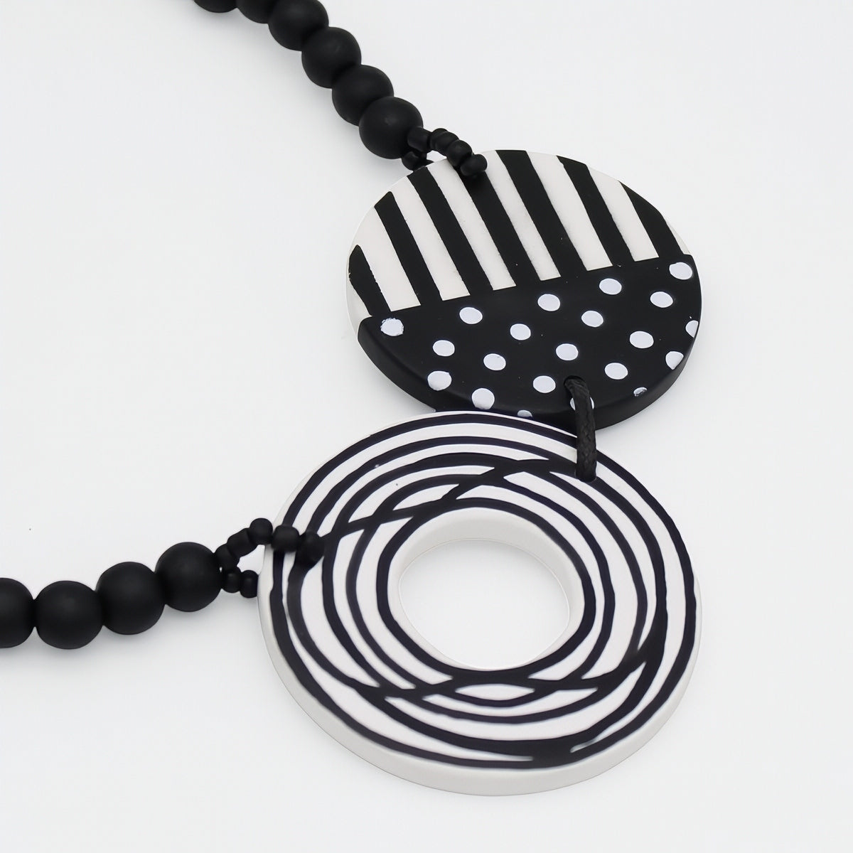 Black and white geometric pendant necklace featuring a striped and polka-dot circular pendant and a coiled ring pendant, suspended over a black beaded chain on a white background.