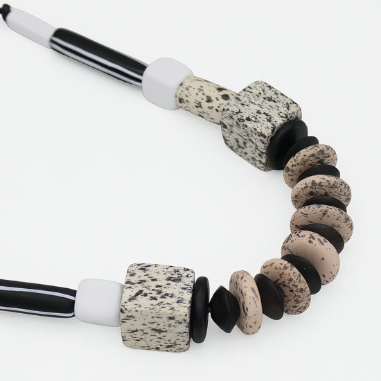 A close-up of a beaded necklace featuring speckled stone beads in beige, black, and white, with striped black and white cord ends, shot against a clean white background.