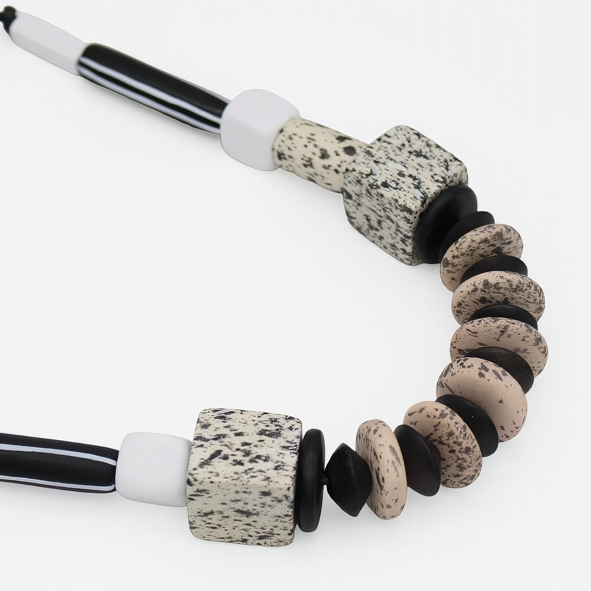 A close-up of a beaded necklace featuring speckled stone beads in beige, black, and white, with striped black and white cord ends, shot against a clean white background.