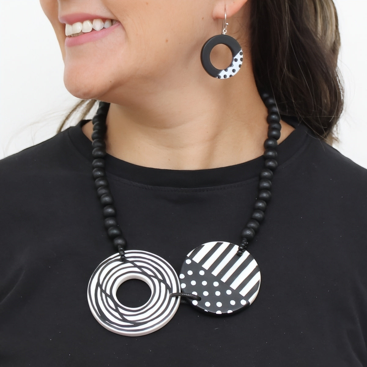 A close-up of a person wearing a black beaded necklace with two large circular pendant pieces featuring black-and-white geometric patterns, along with a matching dangling earring.