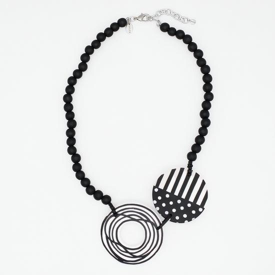 A black beaded statement necklace featuring two circular pendant elements: one with black and white stripes and one with black and white polka dots, displayed on a clean white background.
