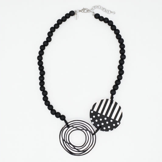 A black beaded statement necklace featuring two circular pendant elements: one with black and white stripes and one with black and white polka dots, displayed on a clean white background.