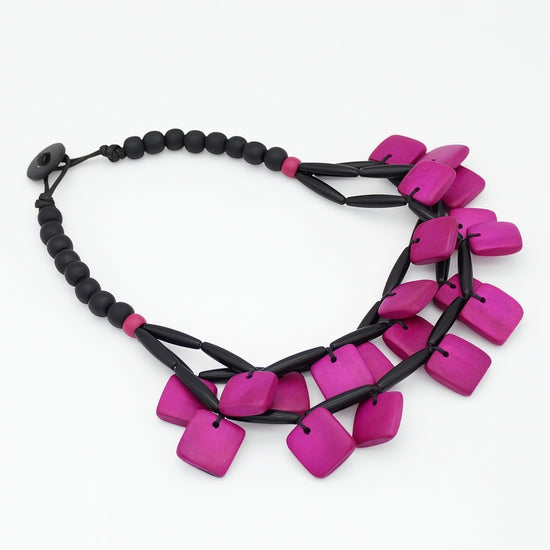 A statement necklace featuring bright magenta-pink square beads and black cylindrical links, displayed on a clean white background.