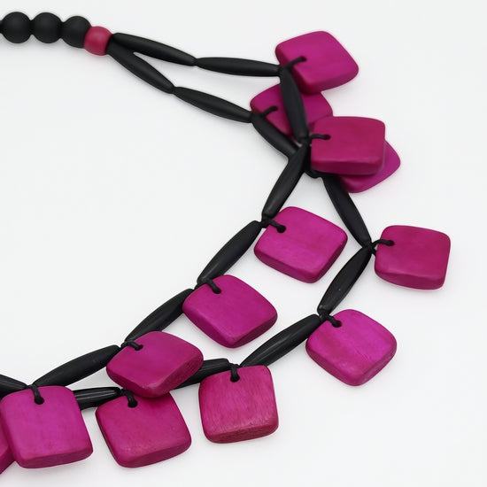 A close-up of a modern statement necklace featuring bright magenta-pink square wooden beads strung on a black cord, set against a plain white background.