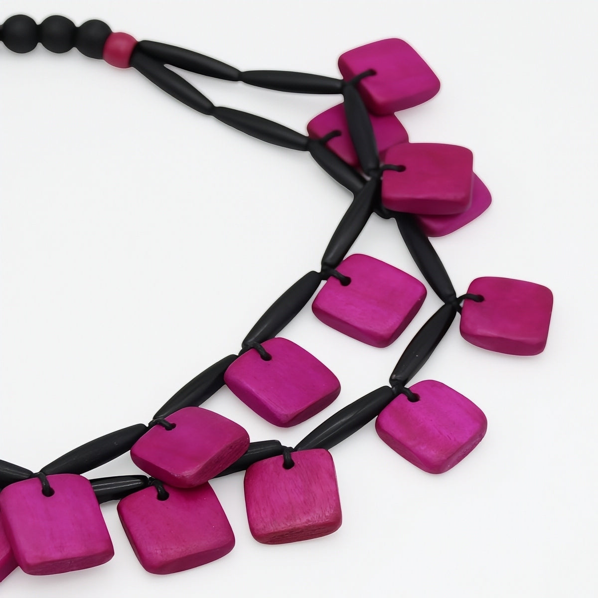 A close-up of a modern statement necklace featuring bright magenta-pink square wooden beads strung on a black cord, set against a plain white background.