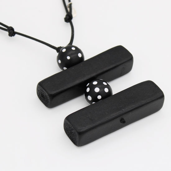 Black wooden pendant necklace with two rectangular bars connected by a black cord and two black beads with white polka dots, shown on a white background.