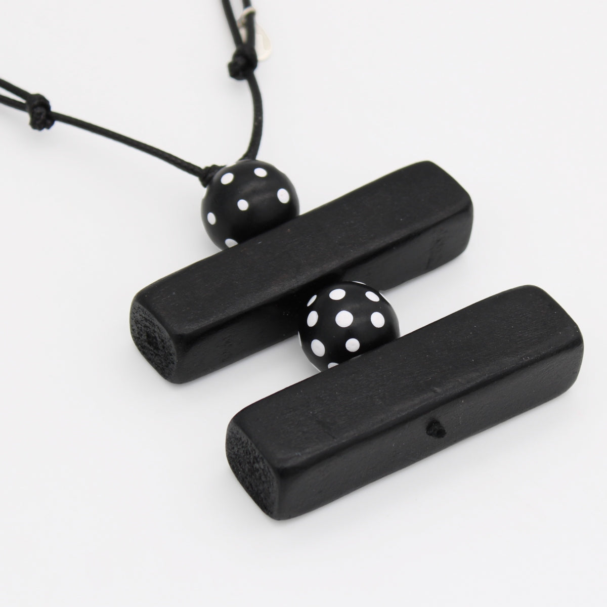 Black wooden pendant necklace with two rectangular bars connected by a black cord and two black beads with white polka dots, shown on a white background.