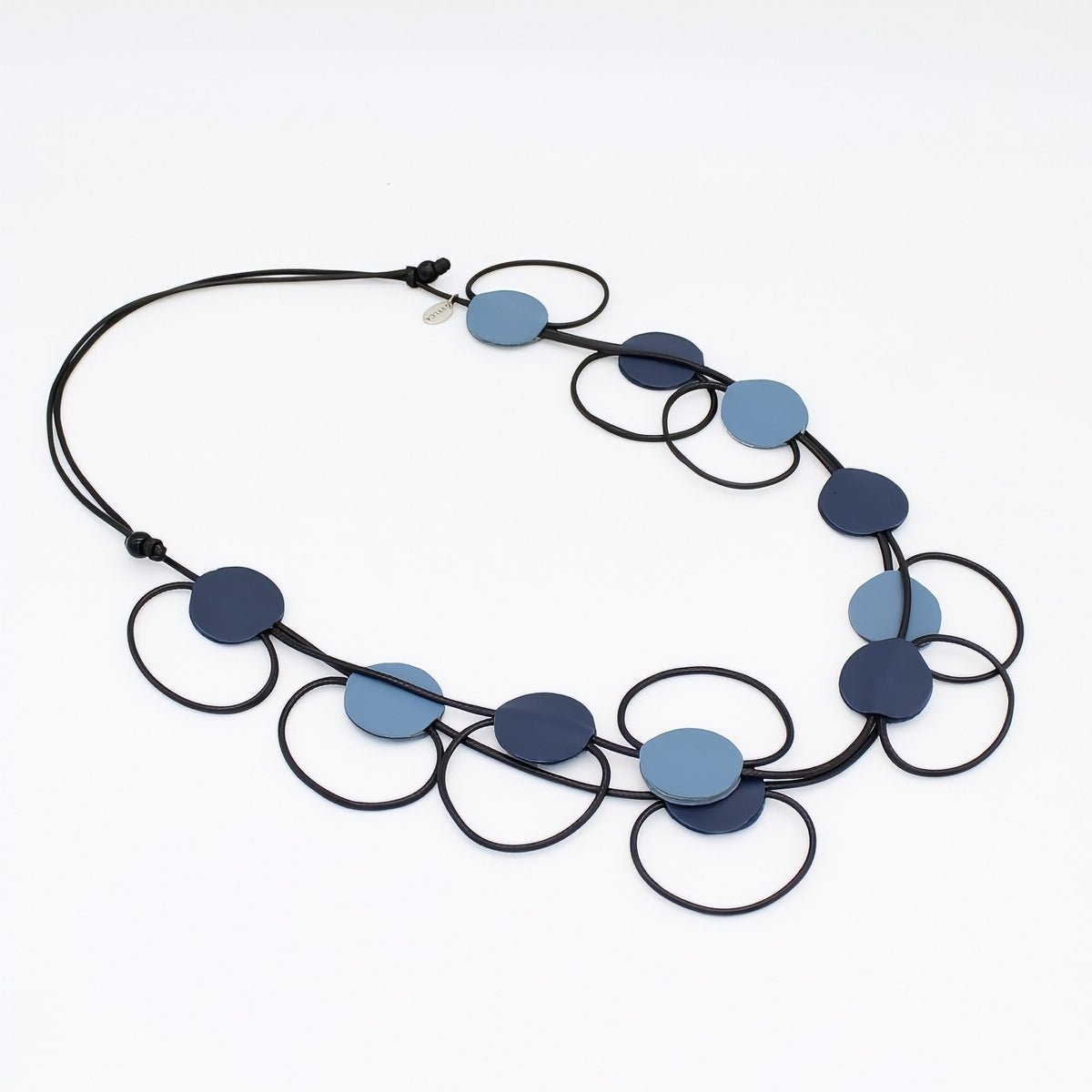 A modern geometric statement necklace featuring flat circular discs in shades of navy and light blue, suspended on a black cord overwire frame, shot against a plain white background.