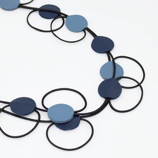 A modern geometric necklace featuring flat circular discs in shades of navy blue, medium blue, and light blue, strung on a black cord against a clean white background.