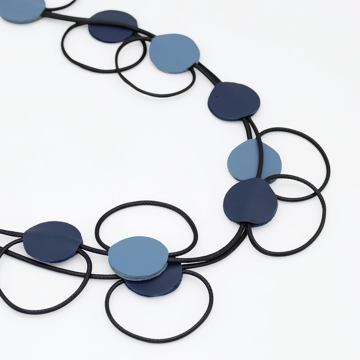 A modern geometric necklace featuring flat circular discs in shades of navy blue, medium blue, and light blue, strung on a black cord against a clean white background.