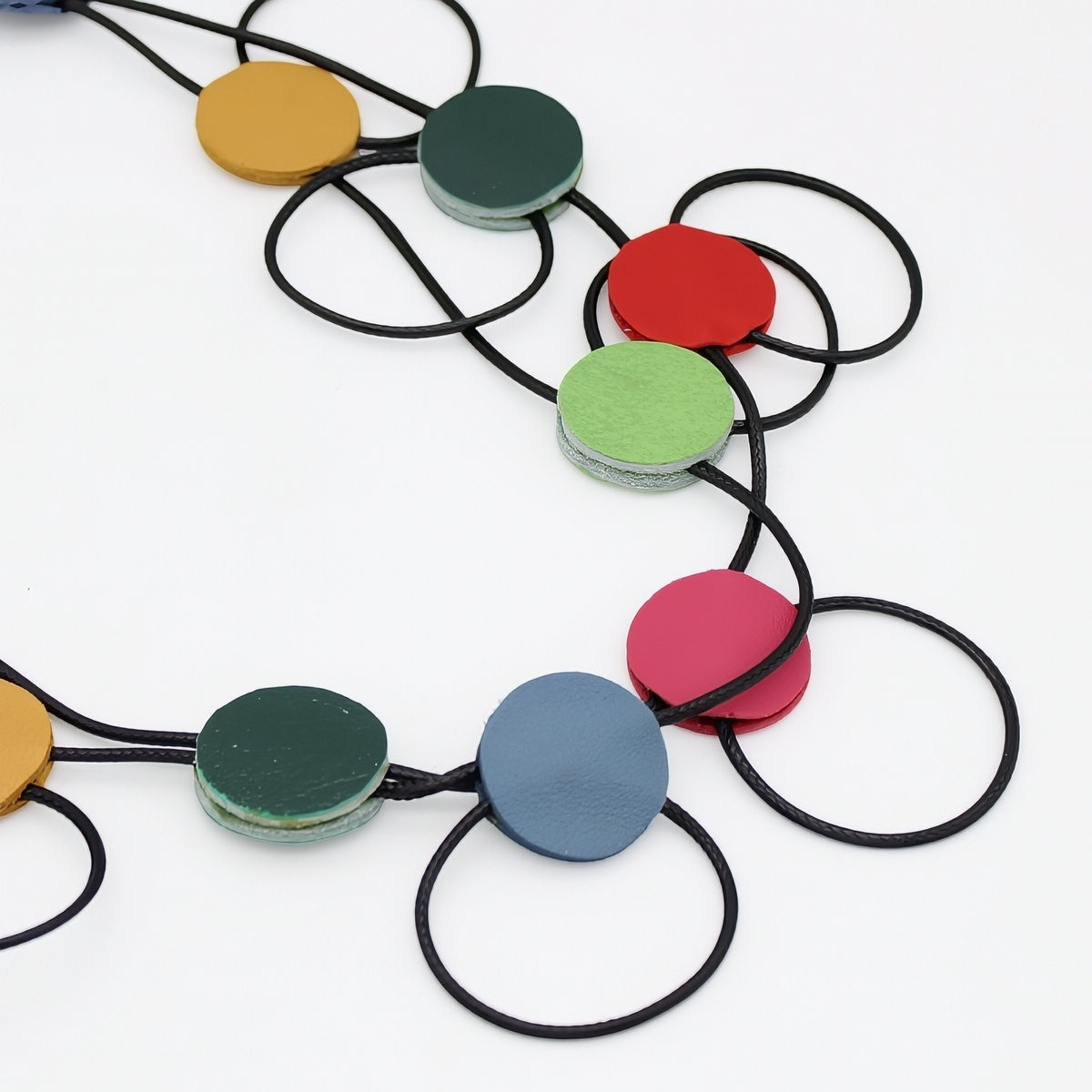 A close-up of a necklace with round wooden disc charms in yellow, green, red, light green, pink, and blue, threaded on black cords against a white background.