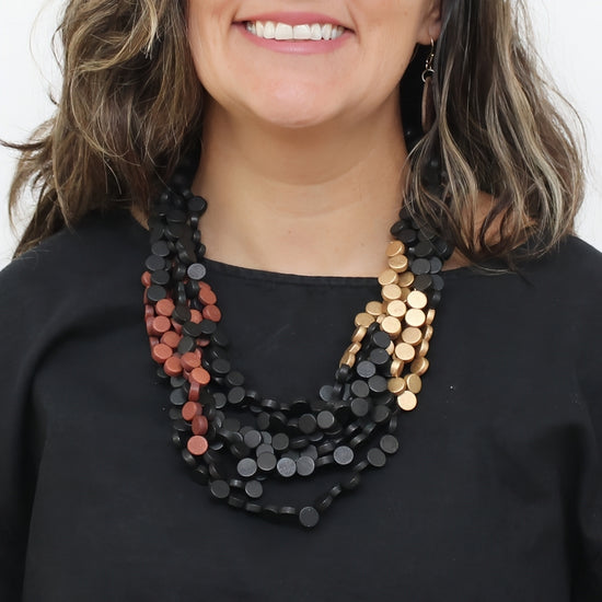 A close-up of a woman wearing a multi-strand statement necklace made of flat circular discs. The necklace features a dominant black strand, a secondary strand of rose gold discs, and a third strand of gold discs, styled over a black top.