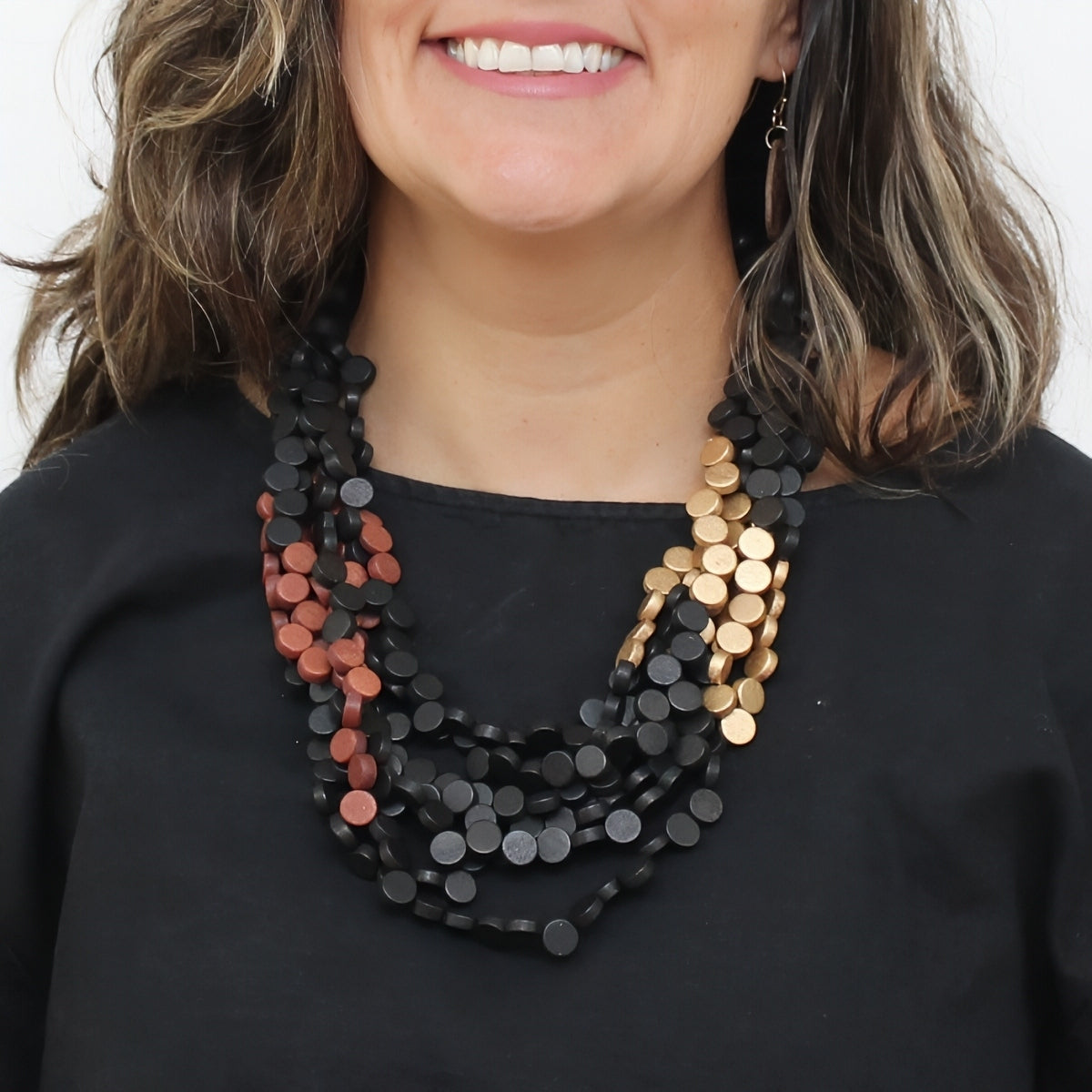 A close-up of a woman wearing a multi-strand statement necklace made of flat circular discs. The necklace features a dominant black strand, a secondary strand of rose gold discs, and a third strand of gold discs, styled over a black top.