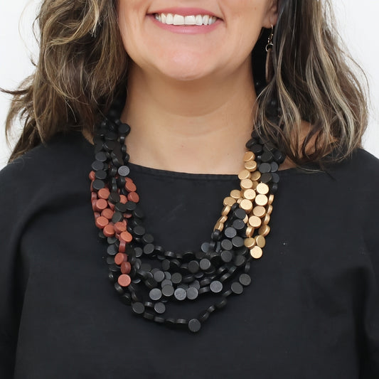 A close-up of a woman wearing a multi-strand statement necklace made of flat circular discs. The necklace features a dominant black strand, a secondary strand of rose gold discs, and a third strand of gold discs, styled over a black top.
