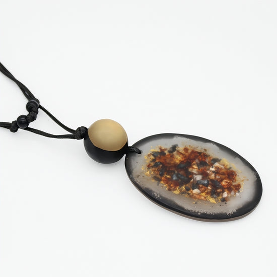 A natural agate geode pendant necklace on a black cord, featuring a polished oval agate slice with a dark rim and colorful crystalline core, accented by a round beige bead on the cord.