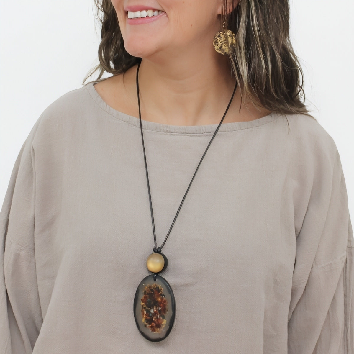 A woman wearing a black cord necklace with a large oval pendant containing dried botanicals, a small round beige bead, and a larger smooth beige bead above it. She is wearing a beige top and gold-toned earrings against a plain white background.