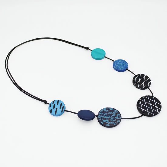 A beaded necklace with a black cord and seven circular and oval beads in shades of blue, teal, and black with geometric patterns, displayed on a clean white background.