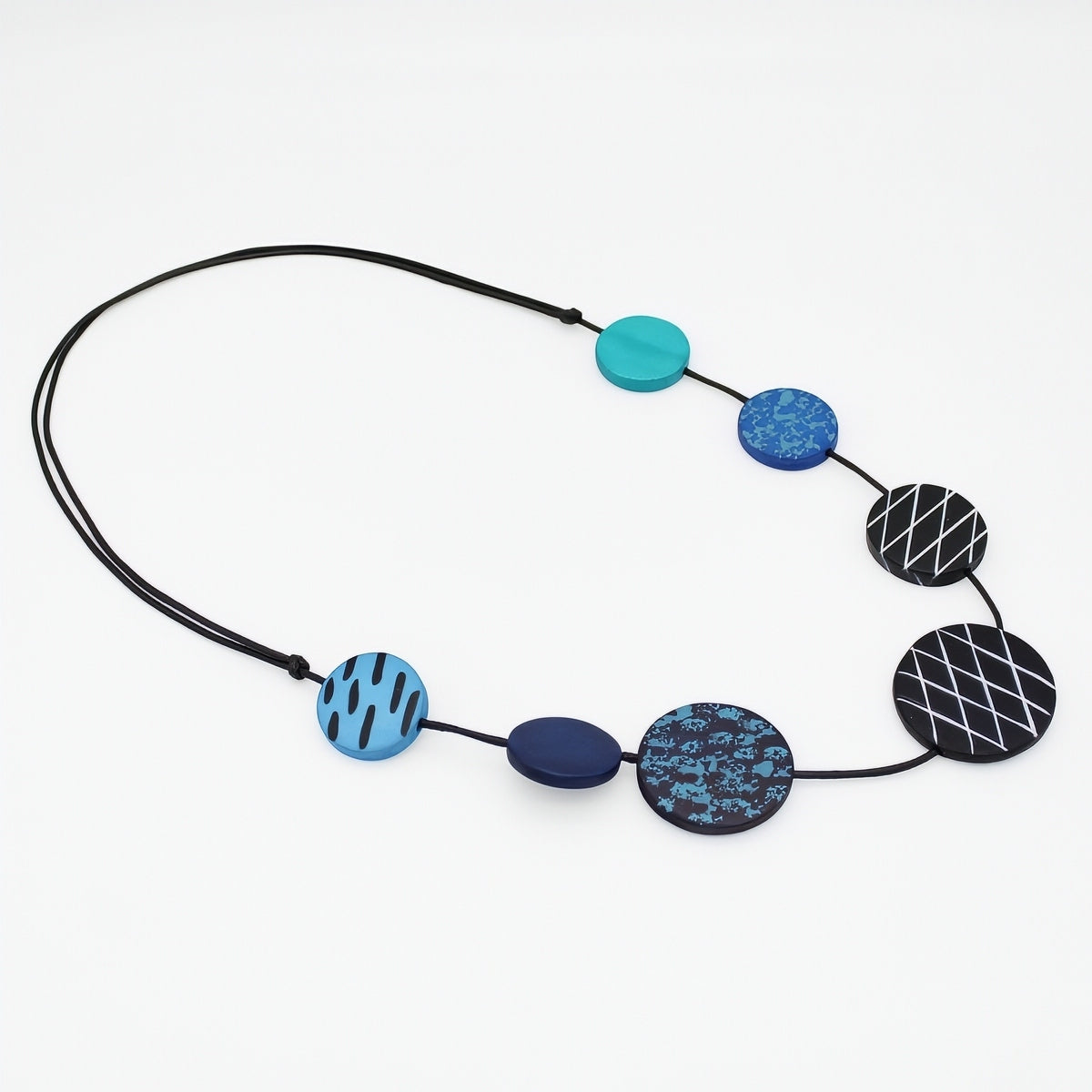 A beaded necklace with a black cord and seven circular and oval beads in shades of blue, teal, and black with geometric patterns, displayed on a clean white background.