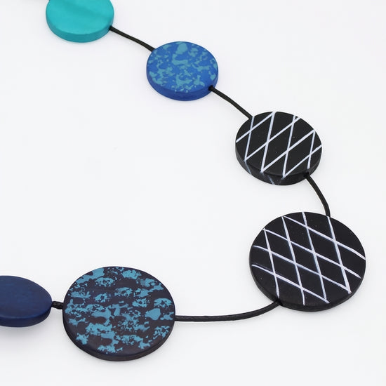 A close-up of a beaded necklace with round and oval beads in turquoise, blue, black, and navy, featuring textured, speckled, and geometric white line patterns, strung on a black cord against a white background.